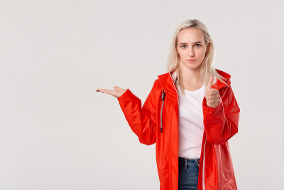 漂亮的金发女孩穿着红色雨衣和兜帽指着空空的手指上一只手孤立在白色的背景。广告的地方