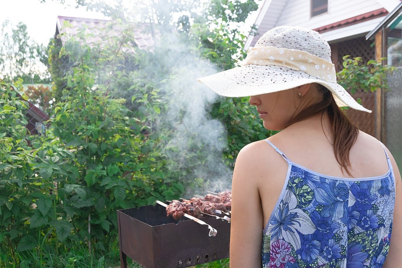 一个戴着草帽的女孩在一个阳光明媚的夏日，在绿叶的映衬下，在烤架上准备着芳香的新鲜肉