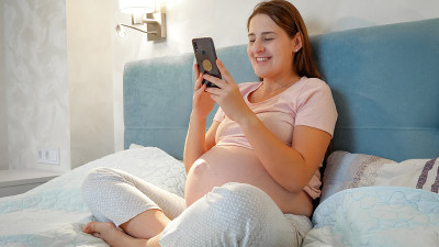 年轻孕妇晚上坐在床上视频通话。怀孕女孩通过智能手机视频会议与朋友或医生交谈。