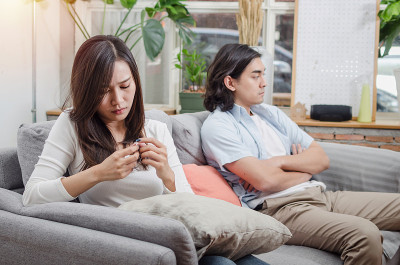 悲伤的年轻亚洲妻子哭着看着她的戒指坐在沙发上后，与丈夫在她身后的房子内部一起争吵，爱，离婚夫妇，家庭