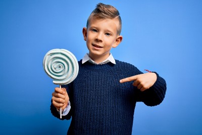 年轻的白人孩子吃糖果棒棒糖在蓝色孤立的背景与惊讶的脸指向自己的手指