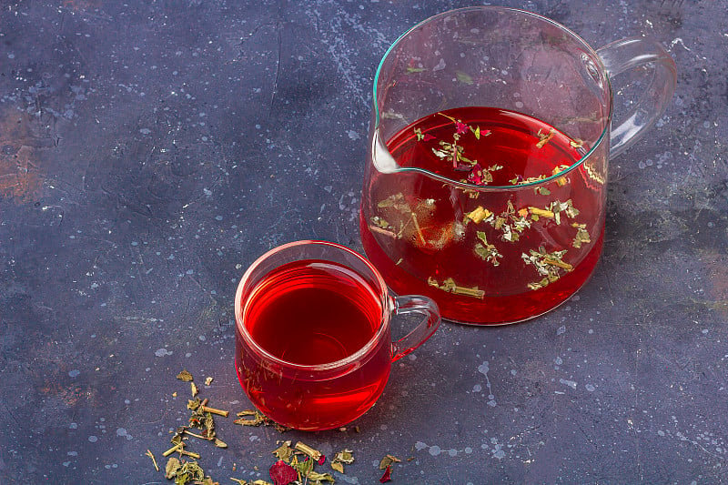 玻璃杯和茶壶中的红茶(芙蓉、木槿、卡卡德)，在干茶叶和花瓣之间，背景为黑色。草药，维生素，治疗感冒和