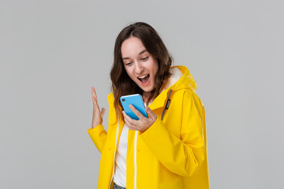 一个穿着黄色雨衣的漂亮的黑发女人在灰色背景下拿着智能手机表达成功的情绪。成功的概念。