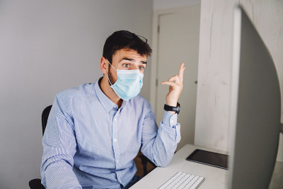 在冠状病毒爆发期间，年轻敬业的商人戴着口罩坐在办公室里使用电脑工作。