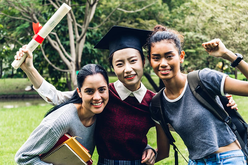 成功的学生年轻女子和学士学位学位毕业生拥抱她的朋友在大学。庆祝毕业和教育理念