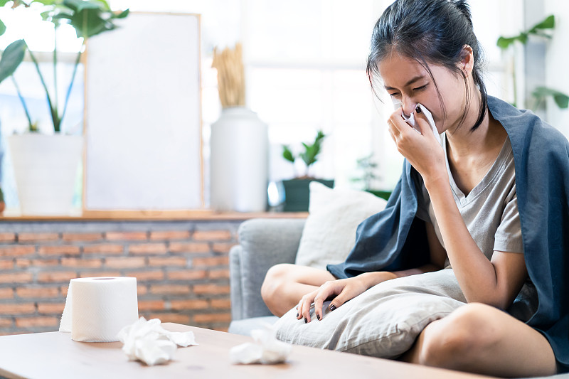 亚洲年轻女子感染冠状病毒。她一个人坐在家里的沙发上。女孩有感冒发热症状，伴有呼吸困难、咳嗽、流鼻涕、