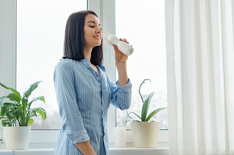 早晨的肖像年轻女子喝牛奶喝酸奶从瓶子，女子站在家里的衬衫附近的窗口，健康的食物吃