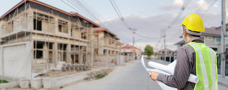年轻的专业工程师戴着防护头盔和图纸纸在房屋建筑工地