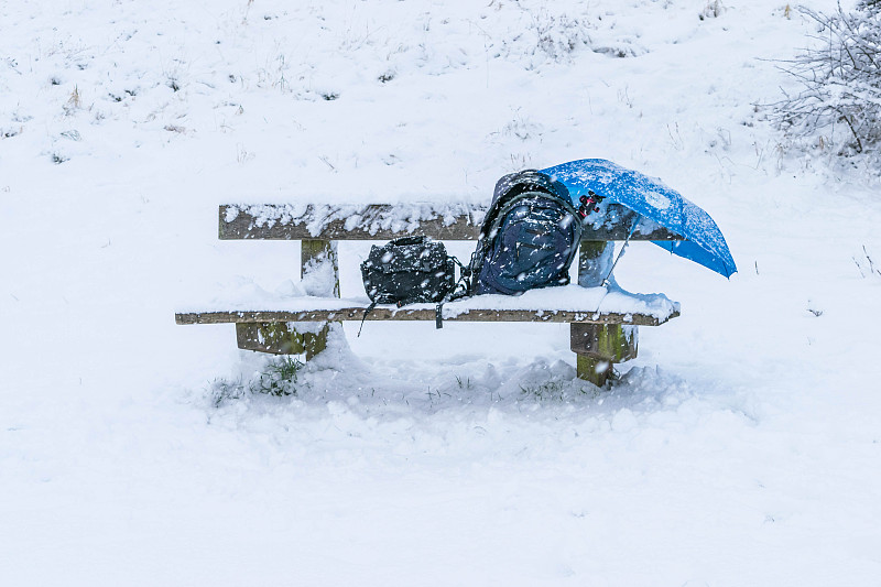 冬季圣诞假期，一场暴风雪过后，公园里的木凳完全被雪覆盖，椅子上有一个背包、雨伞、相机三脚架和一个小包