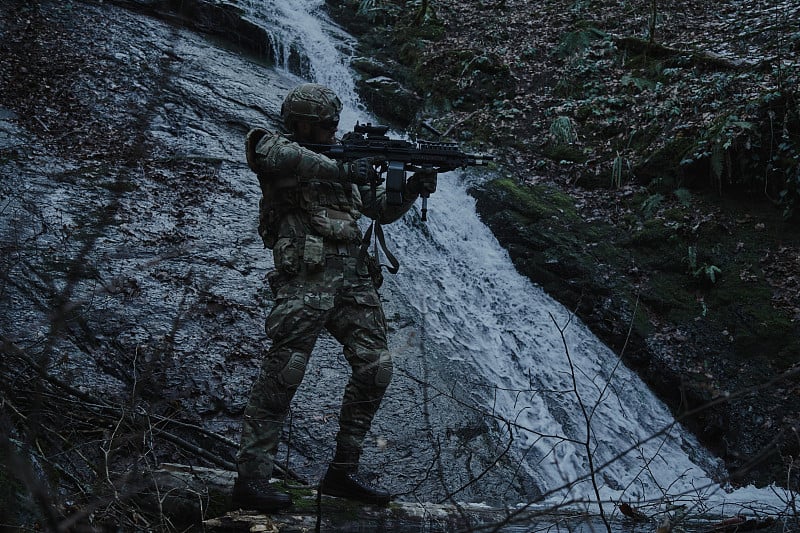 肖像airsoft播放器在专业装备头盔瞄准受害者与枪在森林。在战争中使用武器的士兵