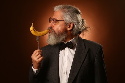画像中是一位白发苍苍的老人，留着蓬乱的小胡子和络腮胡子，穿着白衬衫和灰夹克，戴着眼镜和叉子