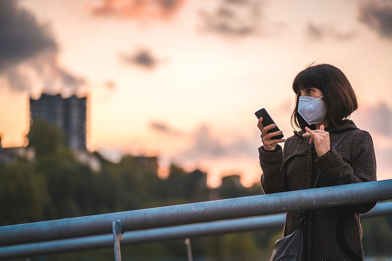 一个戴着面具的黑发姑娘在夕阳下发送音频信息。首次接触未受控制的Covid-19大流行