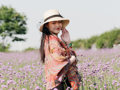身穿黑色马鞭草背心、披着彩色披肩、戴着草帽的美丽女子，在紫色的马鞭草花海中摆姿势，一头乌黑长发的迷人