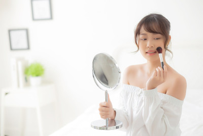 美丽肖像年轻的亚洲女人微笑着看着脸镜子在卧室用刷脸颊化妆，美丽的女孩拿着腮红，皮肤护理和化妆的概念。