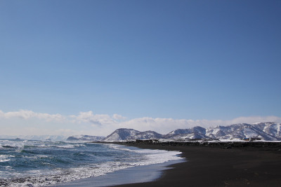 美丽的冬季太平洋海景与大波浪，黑色的火山沙和雪山背景。堪察加半岛阿瓦查湾附近的城市petroplav