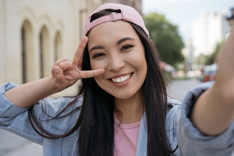 美丽的亚洲女人用手机自拍，显示胜利的标志，享受户外的乐趣。年轻微笑的网红在线视频交流