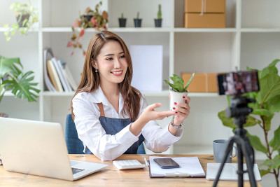 美丽的年轻亚洲商人女人记录视频博客谈话的相机在网上卖植物。