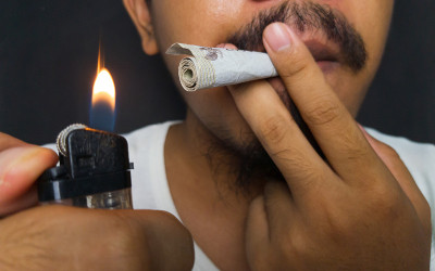 一名留着胡子的亚洲男子身穿白衬衫，在黑色背景前假装吸烟，点燃用印尼盾卷制成的香烟