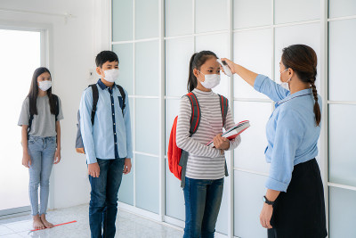 三个亚洲学生戴着口罩站在离他人6英尺远的地方，当学生返回学校时，老师用温度计检查学生的体温。