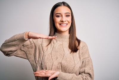 年轻美丽的黑发女人穿着休闲毛衣站在白色的背景手势与手显示大尺寸标志，测量符号。看着镜头微笑。测量的概