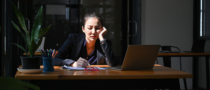 一位疲惫的年轻女商人正拿着笔记本电脑坐在办公桌旁。