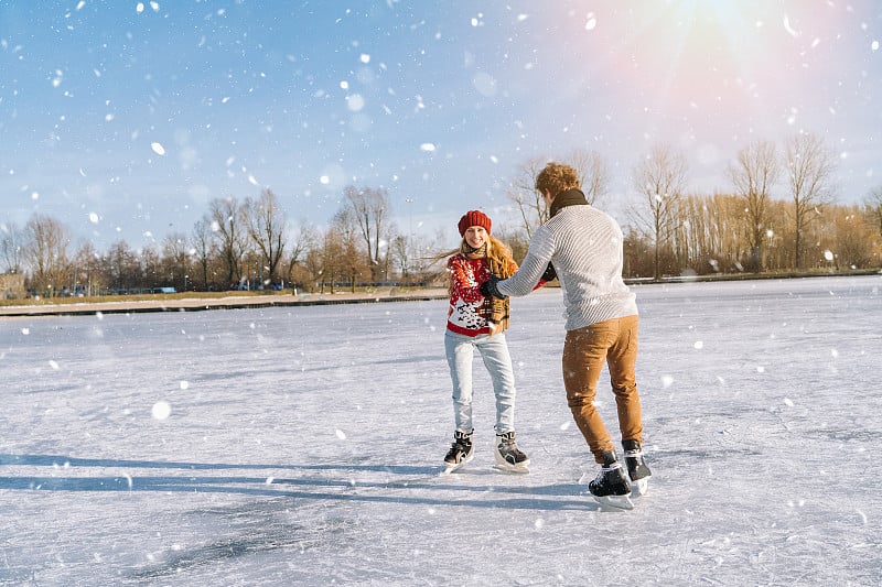 穿着温暖毛衣的情侣在冰上玩耍。女人和男人在阳光明媚的下雪天在户外滑冰。冬季平安夜在冰上竞技场上的活跃