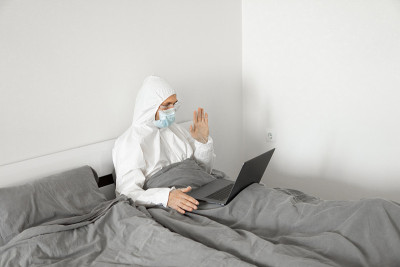 由于冠状病毒疫情，一名身穿白色防护服、戴着医用口罩的男子在家床上工作，并通过网络摄像头向朋友或商业伙