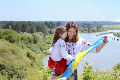 快乐微笑的乌克兰妈妈和女儿穿着vyshyvanka(绣花衬衫)和一个黄蓝色的旗帜，背景是山脉和河流。