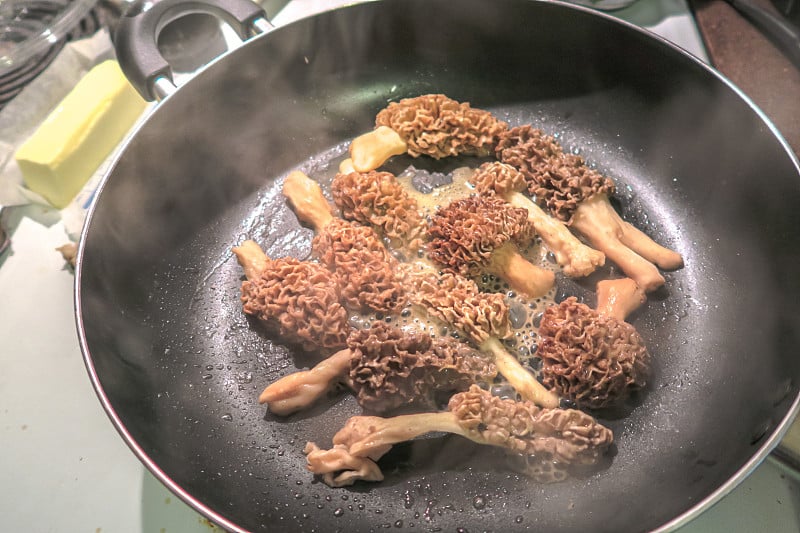 近距离的食物照片，一个煎锅热气腾腾的野生羊肚菌切成两半，涂上黄油在厨房炉子上发出咝咝声。