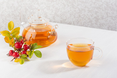 一杯用茶壶和秋熟狗玫瑰枝的玫瑰果茶放在空白的白木桌上。