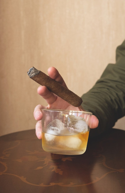 一个穿绿色衬衫的男人拿着一根古巴雪茄和一杯加冰的威士忌。吸烟雪茄的特写镜头。