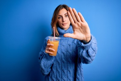 年轻美丽的黑发女人喝健康的橙汁在蓝色的背景与开放的手做停止标志与严肃和自信的表情，防御手势