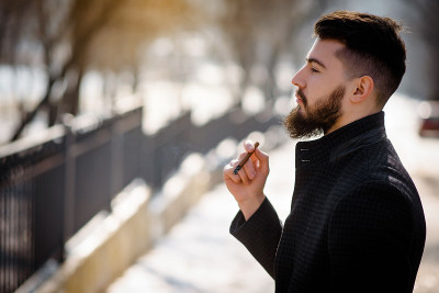 一幅穿着黑色外套、蓄着胡须、英俊潇洒的年轻男子在外面抽烟的肖像。