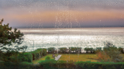 雨点落在一个大的全景窗口望向大海在一个暴风雨，多云的一天，阳光短暂击中大海使它看起来银色。