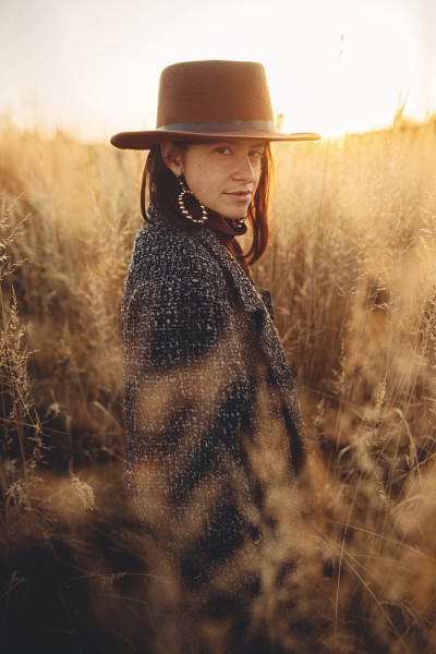 美丽时尚的女人在帽子和复古大衣站在草地在温暖的夕阳光在秋天的田野。宁静的大气。在乡村享受夜晚的时尚年