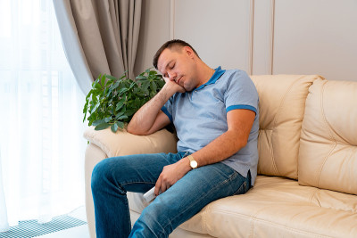 帅哥睡在家里的沙发上，侧影。疲惫的千禧一代睡在客厅的沙发上。男人边看电视边睡觉