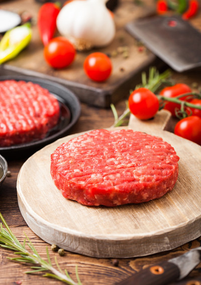 新鲜的生剁碎的自制农民烤牛肉汉堡在圆切板和煎锅与香料和香草和肉斧在木板上。油，大蒜和西红柿。