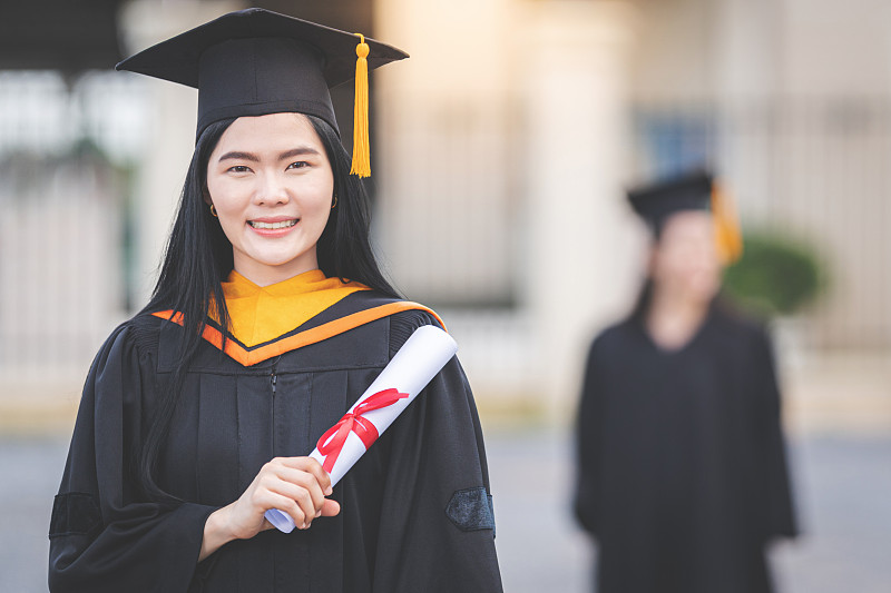 一位年轻的亚洲女子大学毕业生穿着毕业礼服和学士帽，在参加大学毕业典礼后，手持学位证书站在大学大楼前