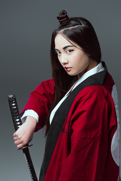 年轻的亚洲妇女在和服持有武士刀，看着孤立的灰色相机