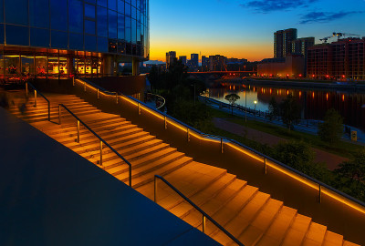 近景的现代摩天大楼照明在晚上，照明楼梯在城市和河流的背景