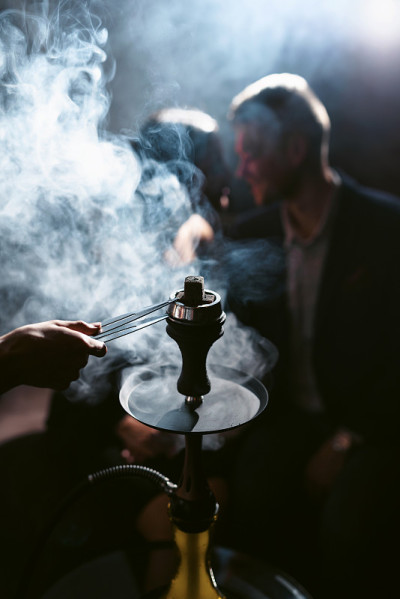 水烟休息室的概念。一对在背光下抽烟喝鸡尾酒的漂亮夫妇。手持钳子的水烟工和卡劳德的手在烟雾缭绕的前景中