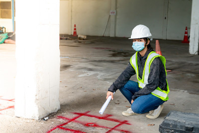 年轻的亚洲女建筑工程师戴着医用口罩遮住嘴，戴着白色安全帽，用卷尺丈量地面，标出施工区域。