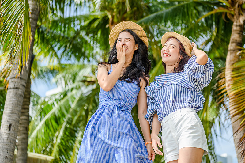 两个快乐的女人拿着草帽在椰子树的海滩上