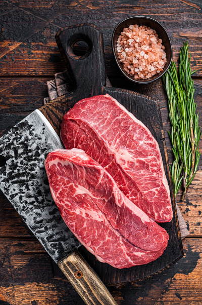 生的生肩刃或平铁牛肉排在木制砧板上与切肉刀。深色木制背景。俯视图