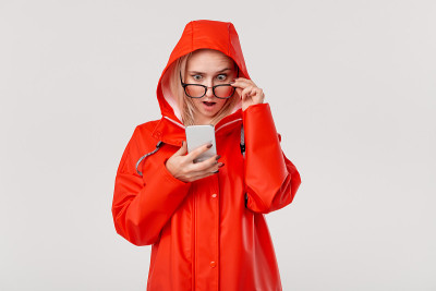 漂亮的金发女人穿着红色雨衣，带着兜帽，看起来惊讶地查看智能手机的通知隔离在白色的背景。在一个寒冷的雨