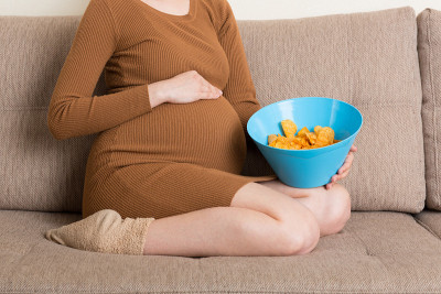 近距离的孕妇坐在沙发上吃薯片从碗在家里。怀孕期间感到饥饿