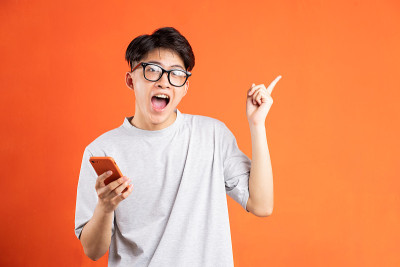 一名年轻的亚洲男子手里拿着智能手机指着前方，孤立在橙色背景下