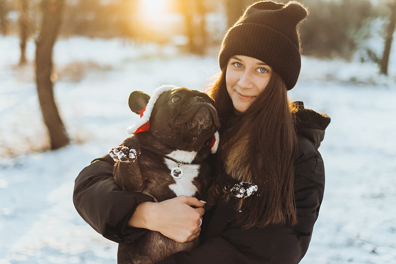 美丽的十几岁的女孩抱着一只法国斗牛犬在她的手臂在冬天。公园里遛狗的女孩