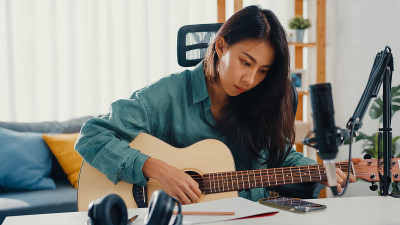 快乐的亚洲女性作曲家弹奏原声吉他，从智能手机上听歌曲，思考，写笔记，在纸上写歌词，在家庭工作室的客厅