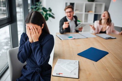 年轻女子被粗鲁的同事伤害。她坐在靠窗的桌旁，双手捂着脸。人们尖叫着嘲笑她。他们的行为不好。